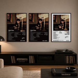 Puede incluir: Tres carteles enmarcados con la portada del álbum "Take Care" de Drake. Cada cartel muestra el título del álbum, la lista de canciones y un retrato de Drake. Los carteles son negros con detalles dorados y se exhiben sobre un mueble de TV negro.