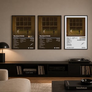 May include: Three framed posters with the text "The Great Divide" and the artist's name, Noah Kahan. The posters feature a window scene with figures and a tracklist. The posters are displayed above a black media console with a record player.