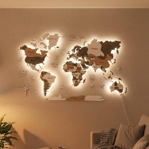 Puede incluir: Un mapa del mundo de madera multicapa para pared en varios tonos de marrón, iluminado por una luz LED trasera de color blanco cálido. El mapa incluye formas de continentes recortadas, nombres de países, iconos de aviones y una rosa de los vientos.