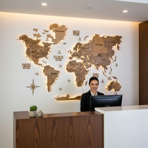May include: A large, multi-layered wooden world map wall art installation featuring backlit illumination. The map displays continents, countries, and ocean names with small wooden airplane and compass icons. It is mounted on a white wall.