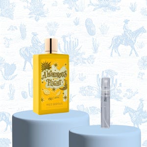 Peut inclure: Un flacon de parfum jaune rectangulaire avec des détails dorés, portant l'inscription "A Wondrous Flight", est présenté sur un socle bleu clair. Un petit vaporisateur transparent est également visible. Le fond est orné d'un motif désertique bleu clair et blanc.