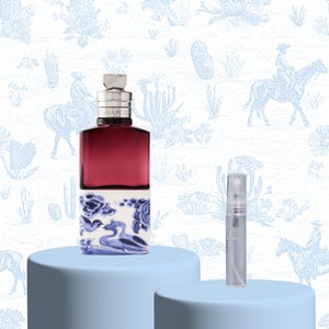 Peut inclure: Un flacon de parfum bordeaux avec un bouchon argenté et un motif de style Delft sur la base, à côté d'un petit vaporisateur d'échantillon. Le fond présente un motif bleu clair avec des motifs de désert et de cow-boy. Le flacon est posé sur un socle bleu clair.