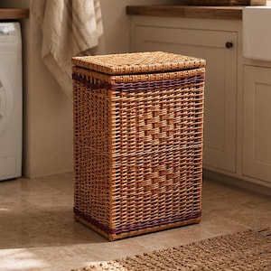May include: A rectangular wicker laundry hamper with a lid. The hamper is woven in a natural brown color with a dark red trim. The hamper is sitting on a tile floor in a laundry room.