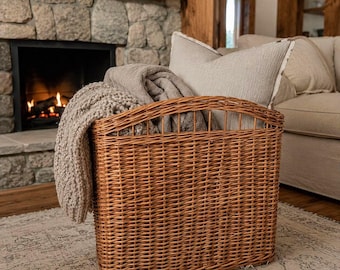 Puede incluir: Una cesta rectangular de mimbre marrón con un patrón tejido, que contiene una manta de punto beige doblada y un cojín gris claro. La cesta presenta un borde superior curvado y está construida con material de caña natural con un tejido visible.