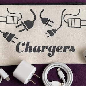 May include: A cream-coloured zipped pouch with the word "Chargers" in black script, and illustrations of electrical plugs. Various white charging cables and power adapters are arranged around the pouch.