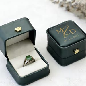 Könnte beinhalten: Zwei dunkelgrüne, quadratische Ringkästchen mit goldenen kronenförmigen Verschlüssen. Ein Kästchen ist geöffnet und zeigt einen silbernen Ring mit grünem Stein auf einem weißen Samtkissen. Das geschlossene Kästchen trägt den Goldaufdruck M&D und 05. 12. 2025.