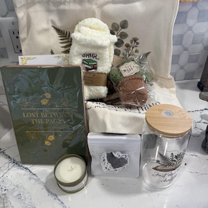 May include: A gift set containing a cream canvas tote bag with botanical prints, a pair of white fuzzy socks with a book graphic, a green book titled Lost Between The Pages, a small candle, a sticker pack, a crochet plant, and a glass tumbler with a bamboo lid.
