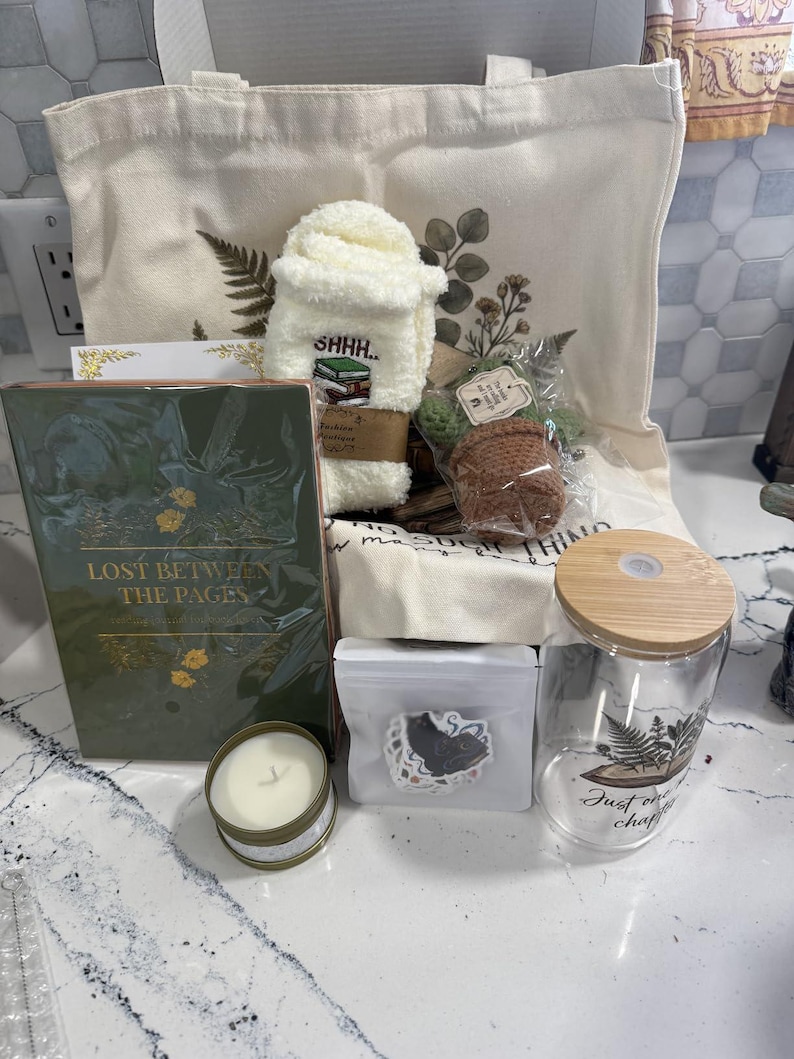 May include: A gift set containing a cream canvas tote bag with botanical prints, a pair of white fuzzy socks with a book graphic, a green book titled Lost Between The Pages, a small candle, a sticker pack, a crochet plant, and a glass tumbler with a bamboo lid.