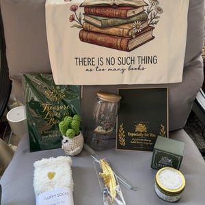 May include: A gift set featuring a cream tote bag with a stack of books and flowers illustration and the text "THERE IS NO SUCH THING as too many books," a green reading journal, a glass jar with a bamboo lid, a crochet cactus, socks, a gold feather bookmark, and a lavender candle.