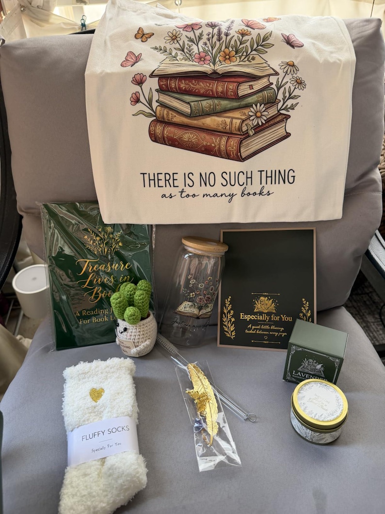 May include: A gift set featuring a cream tote bag with a stack of books and flowers illustration and the text "THERE IS NO SUCH THING as too many books," a green reading journal, a glass jar with a bamboo lid, a crochet cactus, socks, a gold feather bookmark, and a lavender candle.