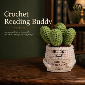 May include: A crocheted cactus in a cream-coloured pot with a smiling face and pink cheeks. The cactus holds a rectangular sign that reads "SHHH... I'M READING" with an open book icon. The item is crafted from yarn with visible textured stitches.