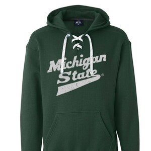 May include: A dark green hooded sweatshirt with a white lace-up detail at the neckline. The sweatshirt has a vintage-style white graphic that says "Michigan State" with a white swoosh underneath.