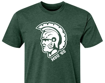 Puede incluir: Una camiseta verde oscuro con un gráfico de cabeza de espartano blanco y el texto "MSU '65".