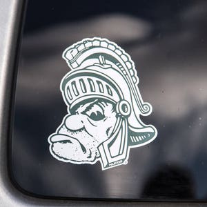 May include: A white and green sticker featuring a stylized Spartan helmet and face. The sticker has a detailed design with a stern expression and is affixed to a dark surface. The sticker has the word "Nudge" on it.