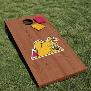 May include: A brown wooden cornhole board with a yellow and red bulldog mascot graphic. The board has two holes and is sitting on green grass. There are two bean bags on the board, one red and one yellow.