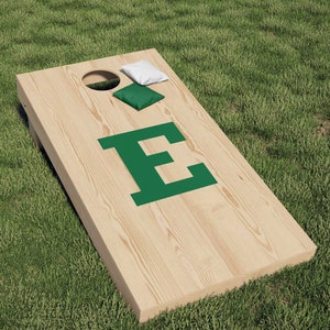 May include: A wooden cornhole game board with a green letter "E" on the front. The board is on a green grassy surface. There are two bean bags, one white and one green, on the board.