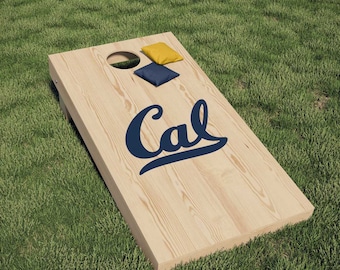 Op de afbeelding: Een houten cornhole-spelbord met het woord "Cal" in blauwe letters. Het bord heeft twee bonenzakken, een gele en een blauwe, erop.