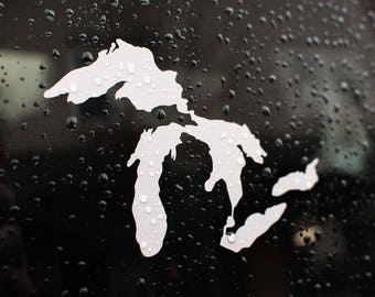 May include: A white vinyl sticker of the Great Lakes on a black background. The sticker is on a car window with raindrops on it.
