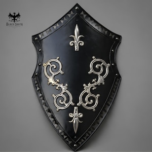 May include: A black heater shield featuring a silver fleur-de-lis at the top and bottom, with symmetrical silver scrollwork in the center. The border is decorated with a repeating triangular pattern and silver rivets. Black Smith Creations logo.