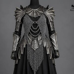 May include: A dark metallic armour set featuring wing-shaped shoulder pauldrons, articulated arm guards, and a chainmail bodice with hanging chain details. The armour includes a gorget and a layered waist piece, all displayed over a black garment.