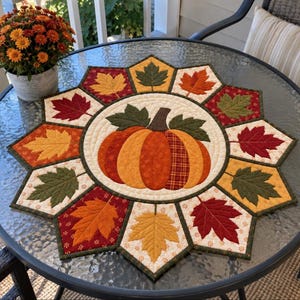 May include: A circular quilted table mat featuring a central orange pumpkin appliqué surrounded by a star-shaped border of twelve fabric leaves in shades of red, orange, yellow, and green, all set against a cream-colored background.