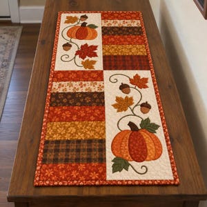 May include: A rectangular quilted table runner featuring a patchwork design of autumn-themed fabrics in shades of orange, brown, and red. The runner includes two cream-colored panels decorated with appliqued orange pumpkins, brown acorns, and leaves.