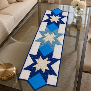 May include: A rectangular quilted table runner featuring a repeating star pattern in shades of navy blue, bright blue, and white. The design incorporates intricate star shapes with gold-toned accents on a white background, finished with a blue border.