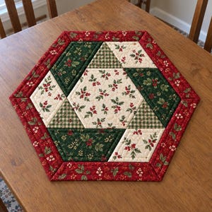 May include: A hexagonal quilted table mat featuring a patchwork design with cream, dark green, and red fabric sections. The pattern includes holly leaf and berry prints, small green gingham checks, and a red border with a floral holly motif.