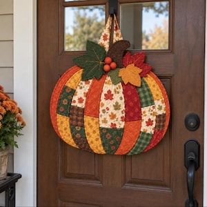 May include: A fabric pumpkin wall hanging featuring a patchwork design of autumn-themed patterns in shades of orange, yellow, green, and brown. The pumpkin is topped with fabric leaves and three small orange spheres, suspended by a matching fabric loop.