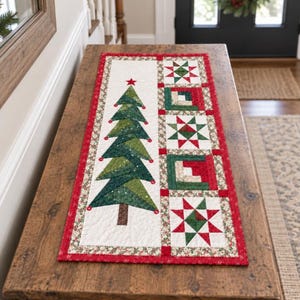 May include: A rectangular quilted table runner featuring a central green fabric Christmas tree with a red star topper on a white background, bordered by a red frame and a vertical strip of alternating star and square geometric quilt blocks.