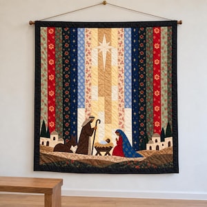 May include: A rectangular wall hanging quilt featuring a nativity scene with a central star, figures in robes, and a donkey. The design uses vertical fabric strips in red, green, blue, and gold patterns, framed by a solid black border and hung by a rod.