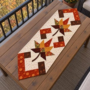May include: A rectangular fabric table runner featuring a patchwork design with two prominent maple leaf motifs. The runner displays a color palette of orange, brown, yellow, and cream, with small floral patterns on the cream-colored fabric sections.