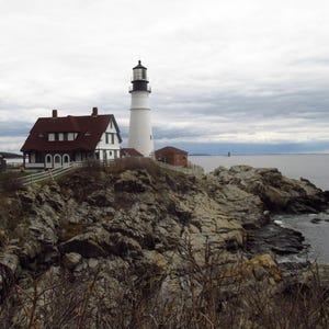 Puede incluir: Escena costera con un faro blanco y negro, un edificio con techo rojo y una pequeña estructura verde. Las estructuras se encuentran en un acantilado rocoso con vistas al océano bajo un cielo nublado. Una valla blanca rodea los edificios.