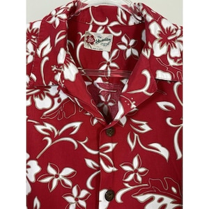 May include: A red short-sleeved button-down shirt featuring a white floral pattern. The shirt has a collar and a label at the neck that reads The Hawaiian Original 2XL. The front closure includes dark brown buttons against the red fabric.