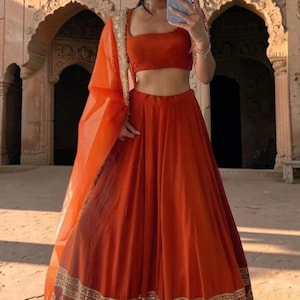 May include: A vibrant orange lehenga set consisting of a sleeveless crop top and a high-waisted floor-length skirt. The skirt features a wide, ornate gold embroidered border with metallic detailing. A matching sheer orange dupatta is draped over one arm.