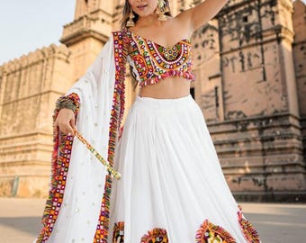 May include: A woman in a white skirt and top with colorful embroidery and fringe. The top is a crop top with a scoop neckline. The skirt has embroidered patches. She is holding a stick in each hand. The background is a stone building.