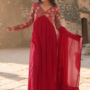 Peut inclure: Longue robe rouge avec un col en V et des manches longues. Le corsage est orné de broderies complexes dorées, rouges et bleues. Une écharpe rouge transparente est drapée sur l'épaule. Parfait pour les occasions spéciales.