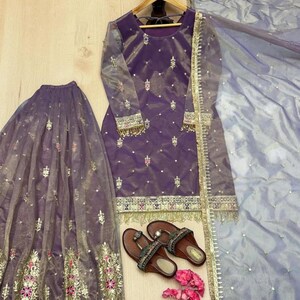 May include: A formal purple outfit with gold embroidery, including a long-sleeved top, skirt, and scarf. Brown sandals and pink flowers are also present. The outfit is displayed on a light wooden surface.