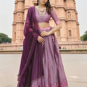 May include: A lavender lehenga choli set featuring a cropped blouse with half-length sleeves and a matching floor-length skirt with a floral border. A deep purple sheer dupatta with a scalloped edge is draped over the shoulder and arm.