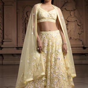 May include: A pale yellow lehenga set featuring a sleeveless V-neck crop top and a floor-length skirt, both adorned with intricate white and pink floral embroidery and silver sequins. A matching sheer yellow dupatta with sequin accents is draped.