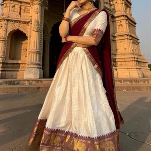 May include: A cream-colored lehenga choli featuring a subtle grid pattern, a deep maroon dupatta with a gold border, and a wide hemline panel decorated with gold elephant motifs. The blouse has elbow-length sleeves and a matching gold border trim.