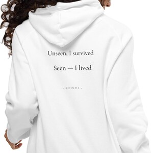 Può includere: Una felpa con cappuccio bianca con testo nero stampato sul retro. Il testo recita: Unseen, I survived. Seen — I lived. Sotto, la parola - S E N T I - è stampata con un carattere più piccolo. La felpa è dotata di maniche lunghe e cappuccio.