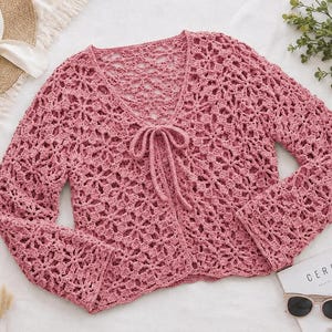 May include: A dusty rose long-sleeve cardigan featuring an intricate openwork crochet pattern throughout. The garment has a V-neckline and a front tie closure made of matching yarn. The sleeves are straight and the hem is finished with a scalloped edge.