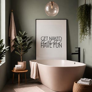 Può includere: Un bagno con una vasca da bagno ovale indipendente rosa chiaro, un poster bianco incorniciato con il testo nero GET NAKED AND HAVE FUN, un tavolino in legno con una pianta, una pianta sospesa, una lampada a sospensione in vetro e asciugamani.