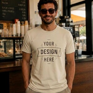 May include: A cream-coloured t-shirt with the text "YOUR DESIGN HERE" in a banner design. The person in the image is wearing sunglasses and standing in a cafe setting.