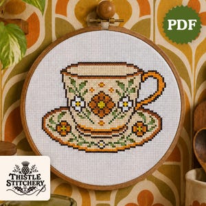 Op de afbeelding: Kruissteekpatroon van een kop en schotel in een houten borduurring. Het ontwerp toont een beige kop met oranje en groene bloemendetails. PDF-pictogram zichtbaar. Thistle Stitchery-logo linksonder.