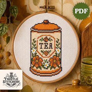 Puede incluir: Un patrón de punto de cruz de un bote de té en un bastidor de bordado. El diseño presenta un recipiente naranja y crema con la palabra "TEA" en el centro, rodeado de flores naranjas y margaritas blancas. El logotipo de Thistle Stitchery está en la esquina inferior izquierda.
