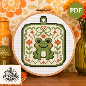May include: A cross-stitch embroidery featuring a green frog design within a square frame, surrounded by floral patterns and set in a wooden hoop. The design includes orange and green accents. The text "PDF" is visible in the upper right corner.