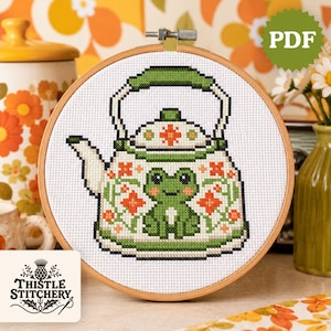 May include: Cross-stitch pattern of a green and white teapot with a frog design, set in a wooden embroidery hoop. The teapot is decorated with orange flowers. The text "PDF" is in a green circle in the upper right corner. The Thistle Stitchery logo is in the lower left corner.