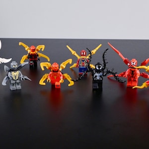 Op de afbeelding: Een verzameling kleurrijke Lego-actiefiguren, met verschillende Spider-Man-personages. De figuren zijn in verschillende kleuren, waaronder rood, blauw, zwart en wit, met kenmerkende spinnenachtige aanhangsels. De figuren zijn gerangschikt op een zwart oppervlak.